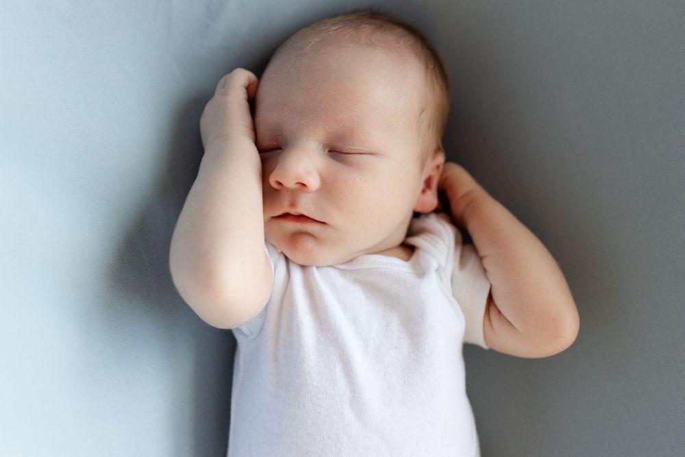 Sleeping newborn in white onesie with hands raised near face on light background.