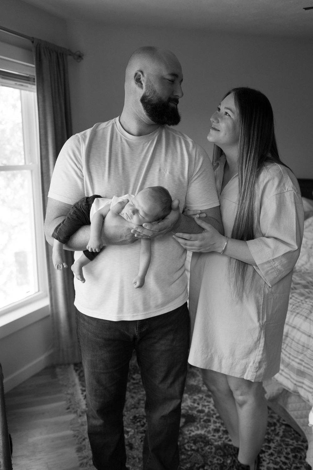 Family of three shares tender moment with newborn by bright window in casual home setting.