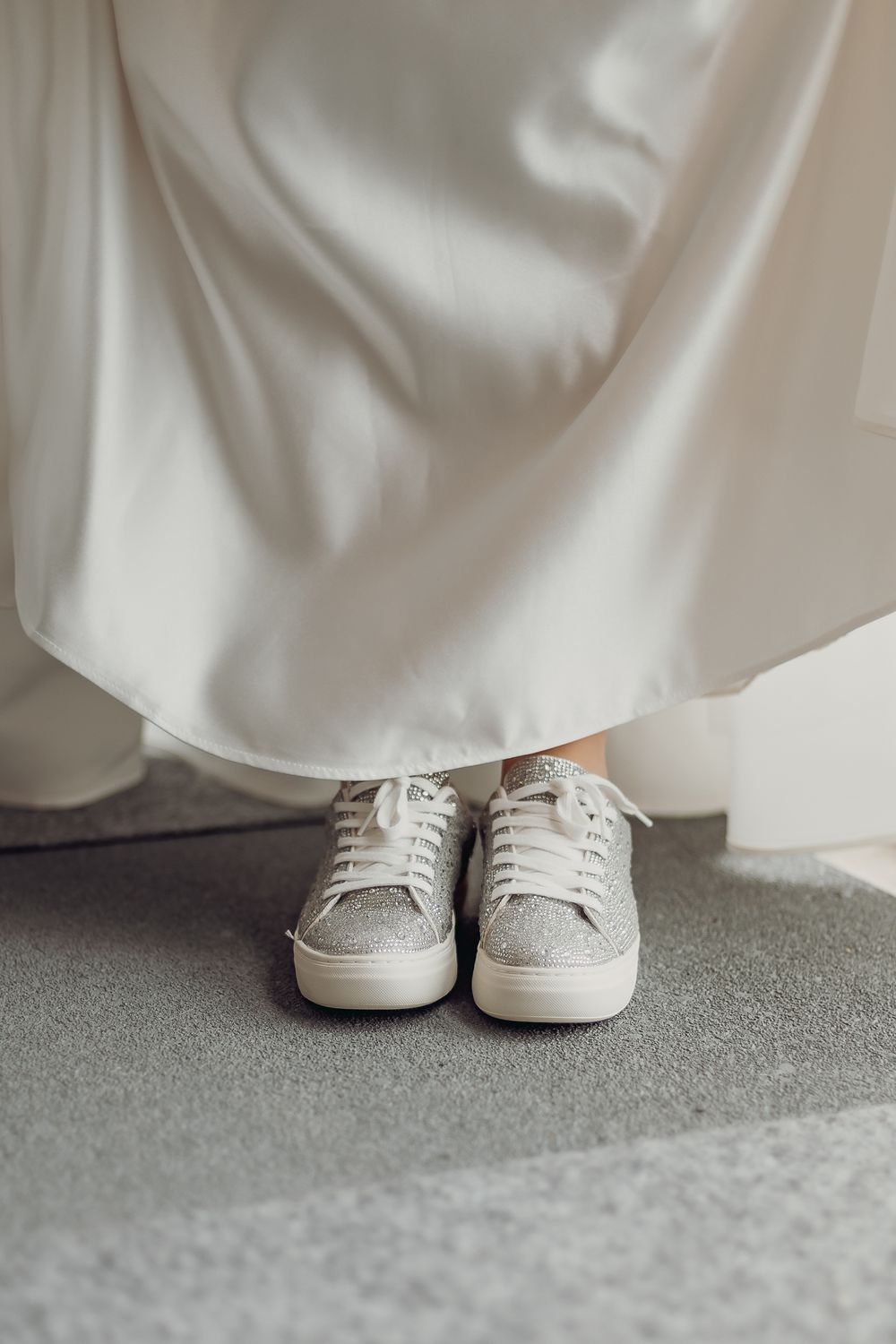 White wedding dress with sparkly sneakers peeking out from underneath on a grey carpet.