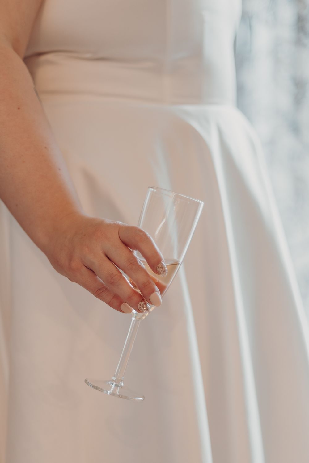 Hand holding champagne glass while wearing elegant white dress.