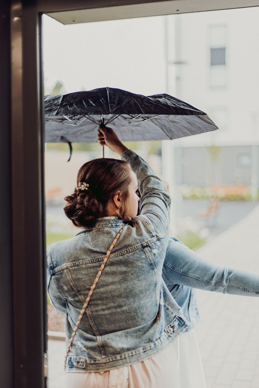 Person in denim jacket holds black umbrella while looking out window.
