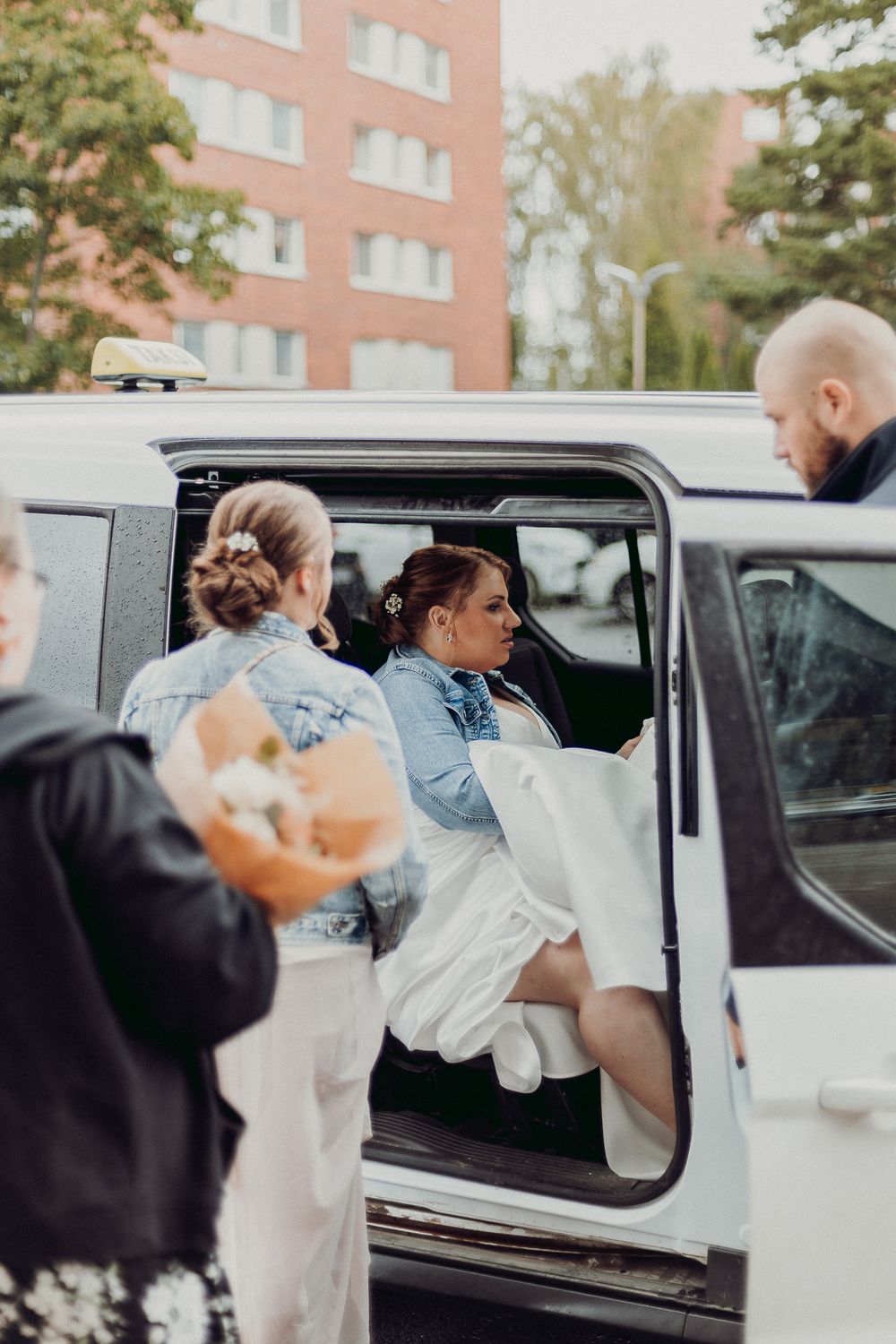 People helping someone in white dress and denim jacket into white car.