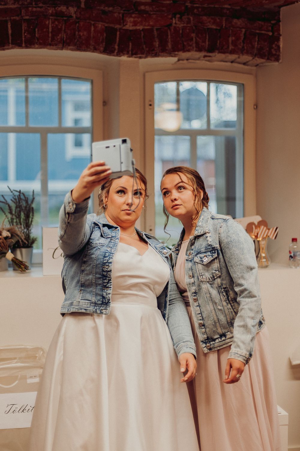 Two people in white dresses and denim jackets posing playfully in a bright room.