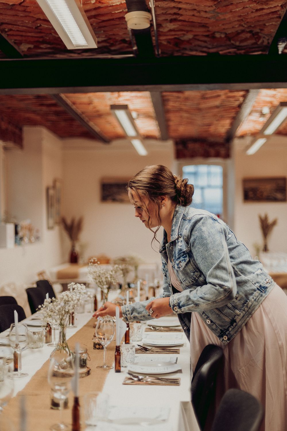 Person in jean jacket and white dress sets elegant table with glassware and place settings.