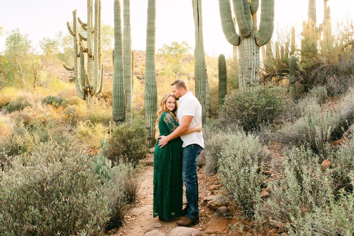 A couple hugging with cactus and greenery around them. She's pregnant and is in a green dress. He's kissing her head and is in a white shirt