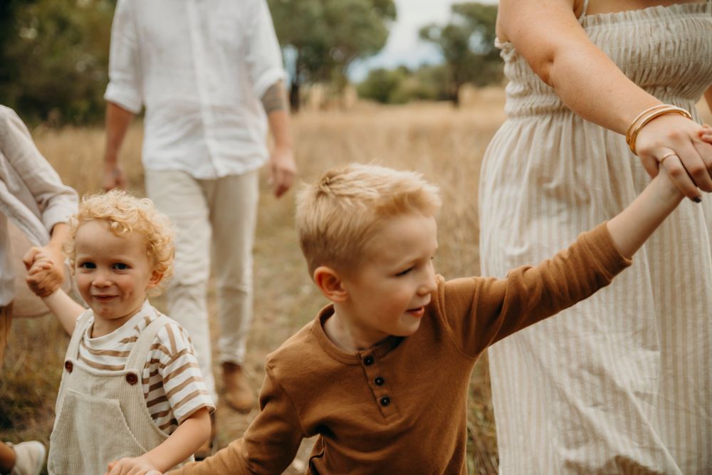 Two young blonde children holding hands walking through golden grass field with parents.