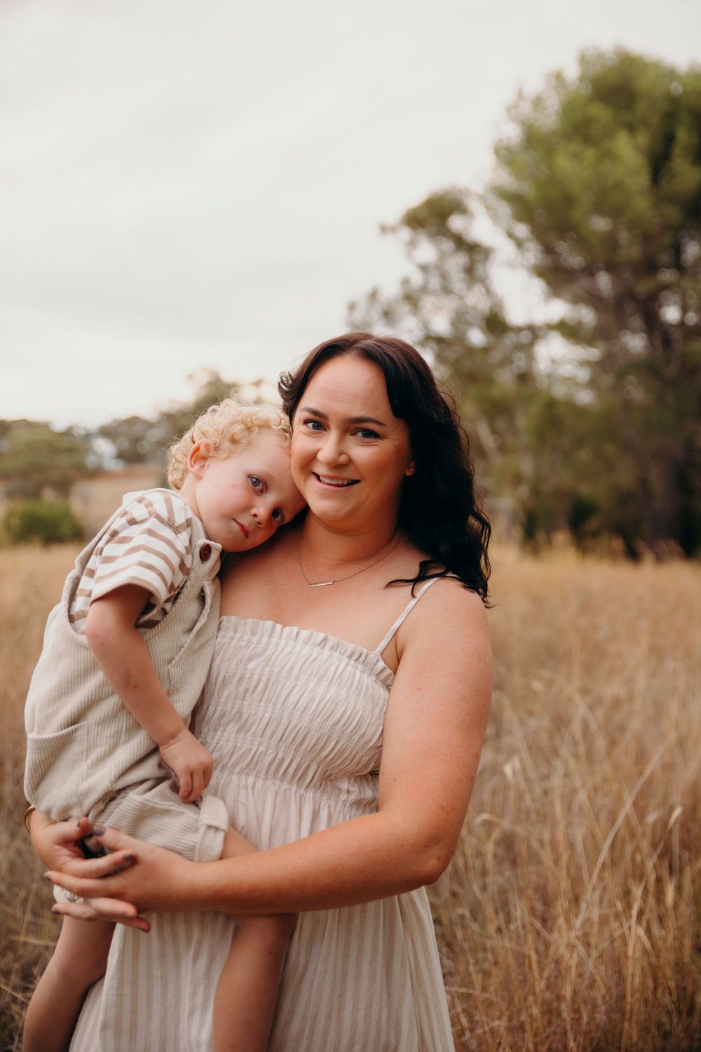 Smiling dark-haired woman in linen dress holding curly-haired toddler in golden grass field.