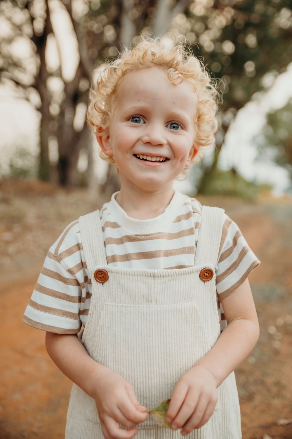 Happy toddler with curly blonde hair wearing striped shirt and overalls smiles outdoors on a dirt path.