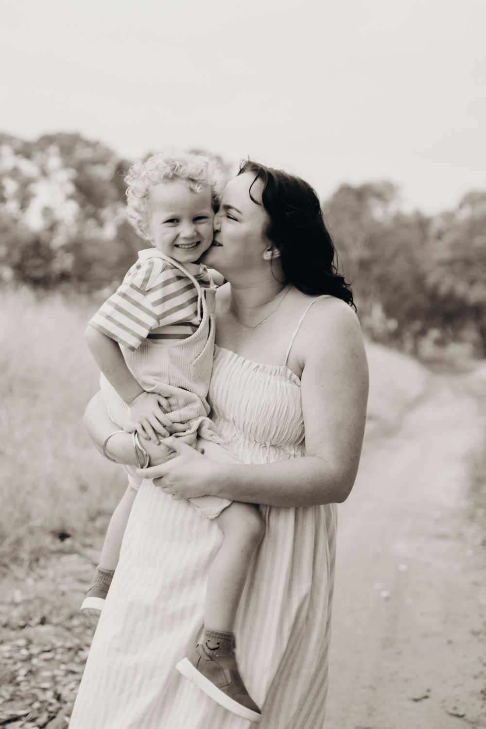Black and white photo of a mother kissing her curly-haired toddler on a rural dirt path.