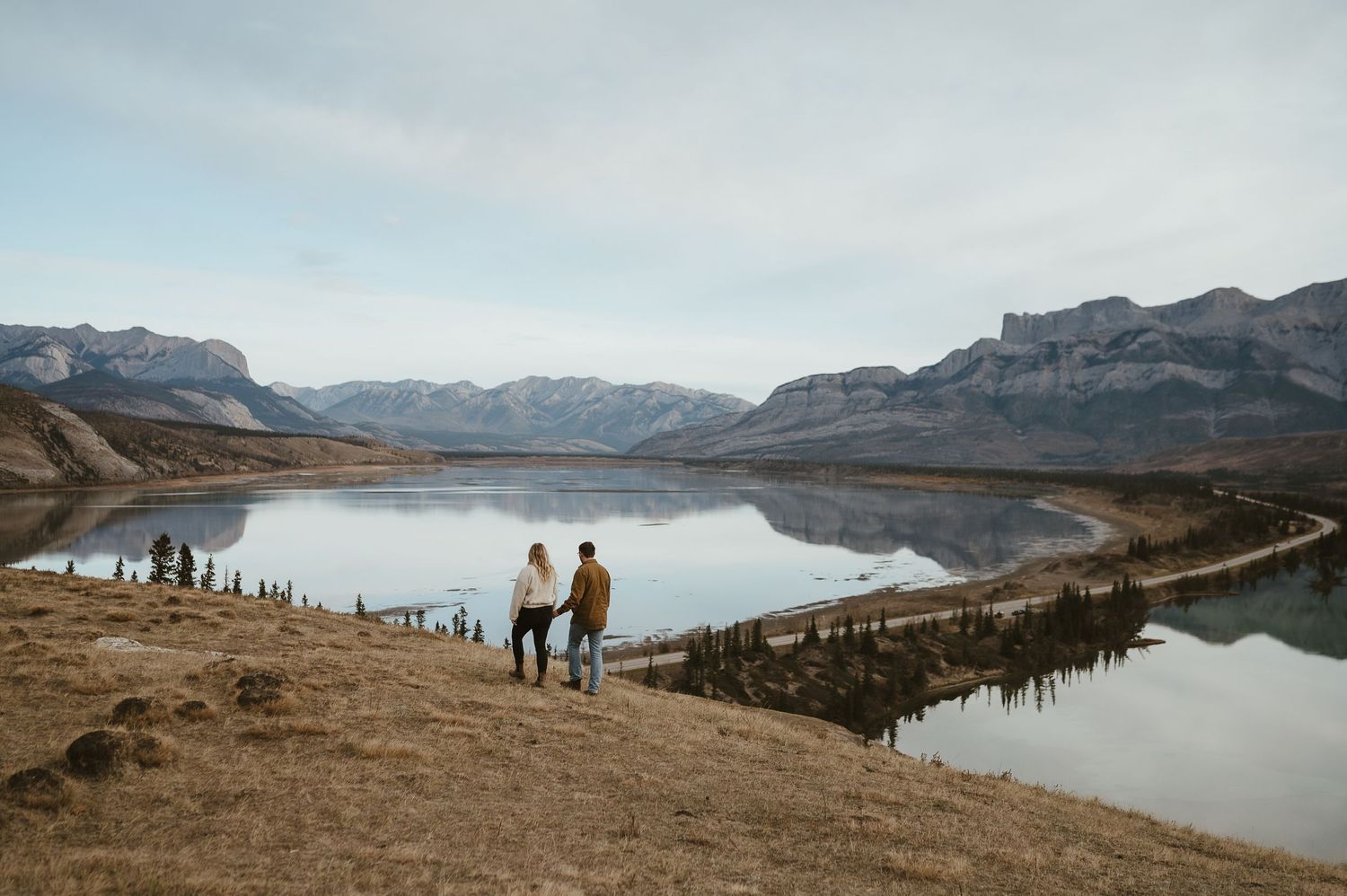 Couple stands on a cliff overlooking a serene mountain lake with dramatic peaks in the background.