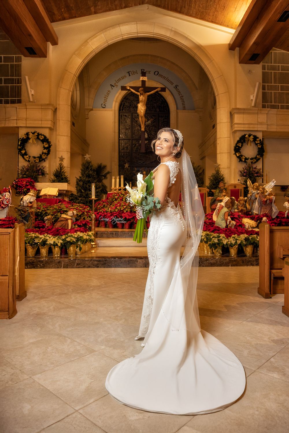 A bride in a white fitted gown with long veil stands in an ornate church decorated with red poinsettias and holiday wreaths.