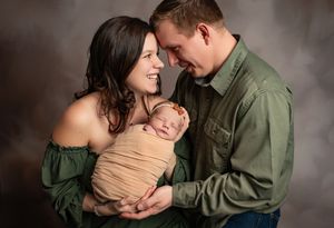 A newborn baby wrapped in tan fabric is held lovingly by parents wearing green tops against a dark backdrop.