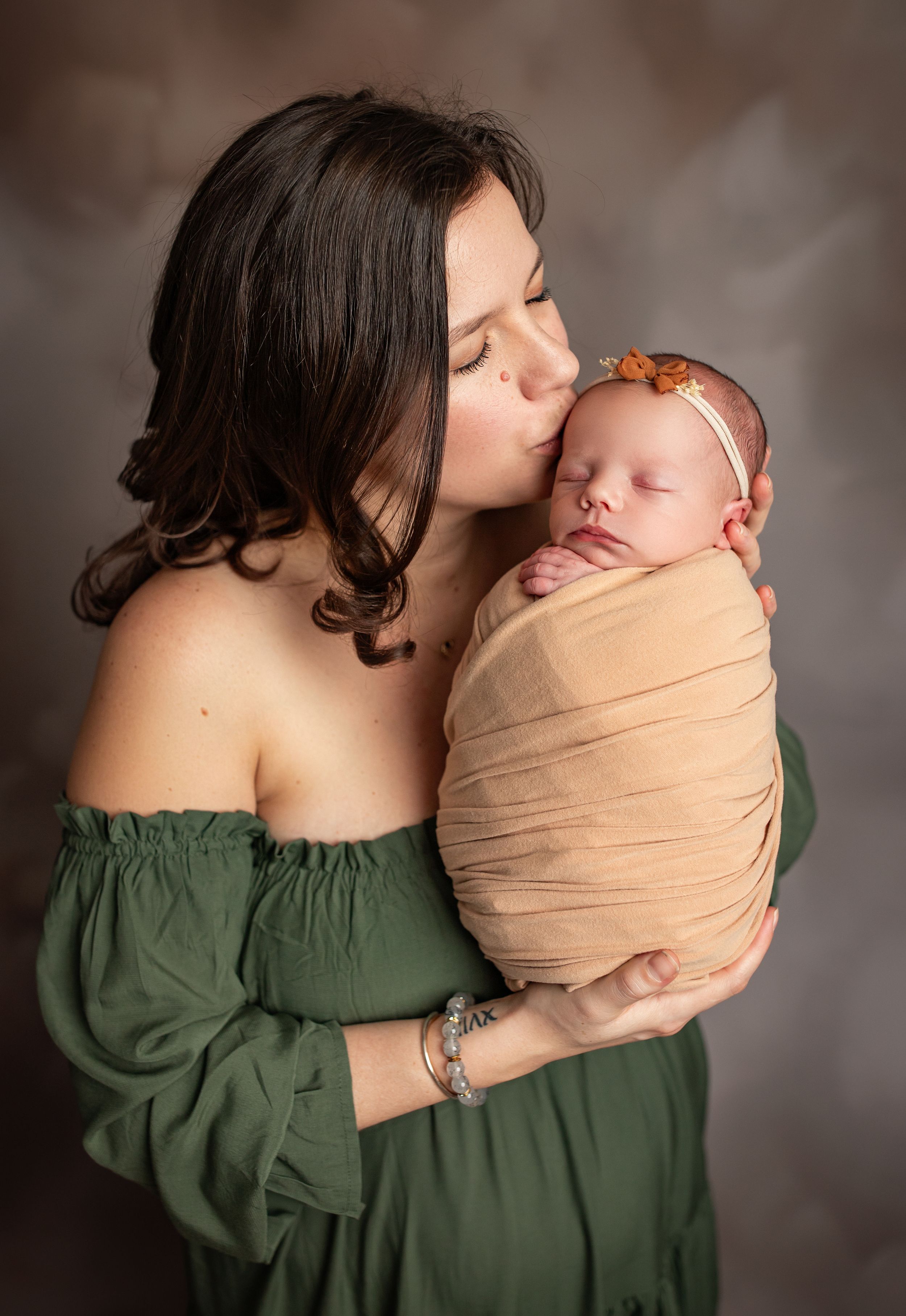 A tender moment as a mother in a green off-shoulder dress kisses her sleeping newborn baby wrapped in beige fabric.
