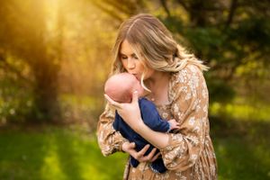 Parent in floral dress holds infant in outdoor sunset setting.
