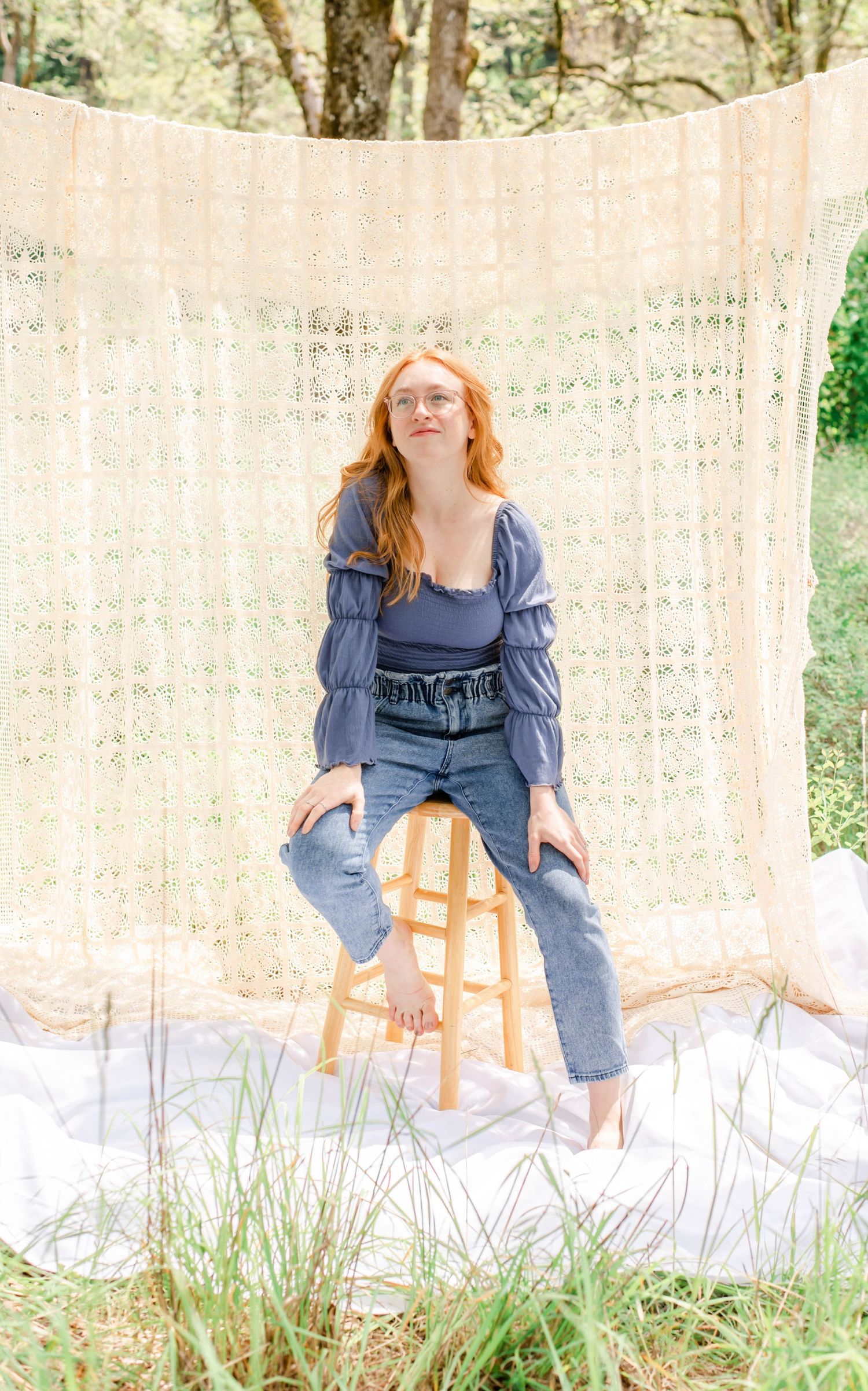 Model in blue peasant top poses expressively on wooden stool with white fabric background.