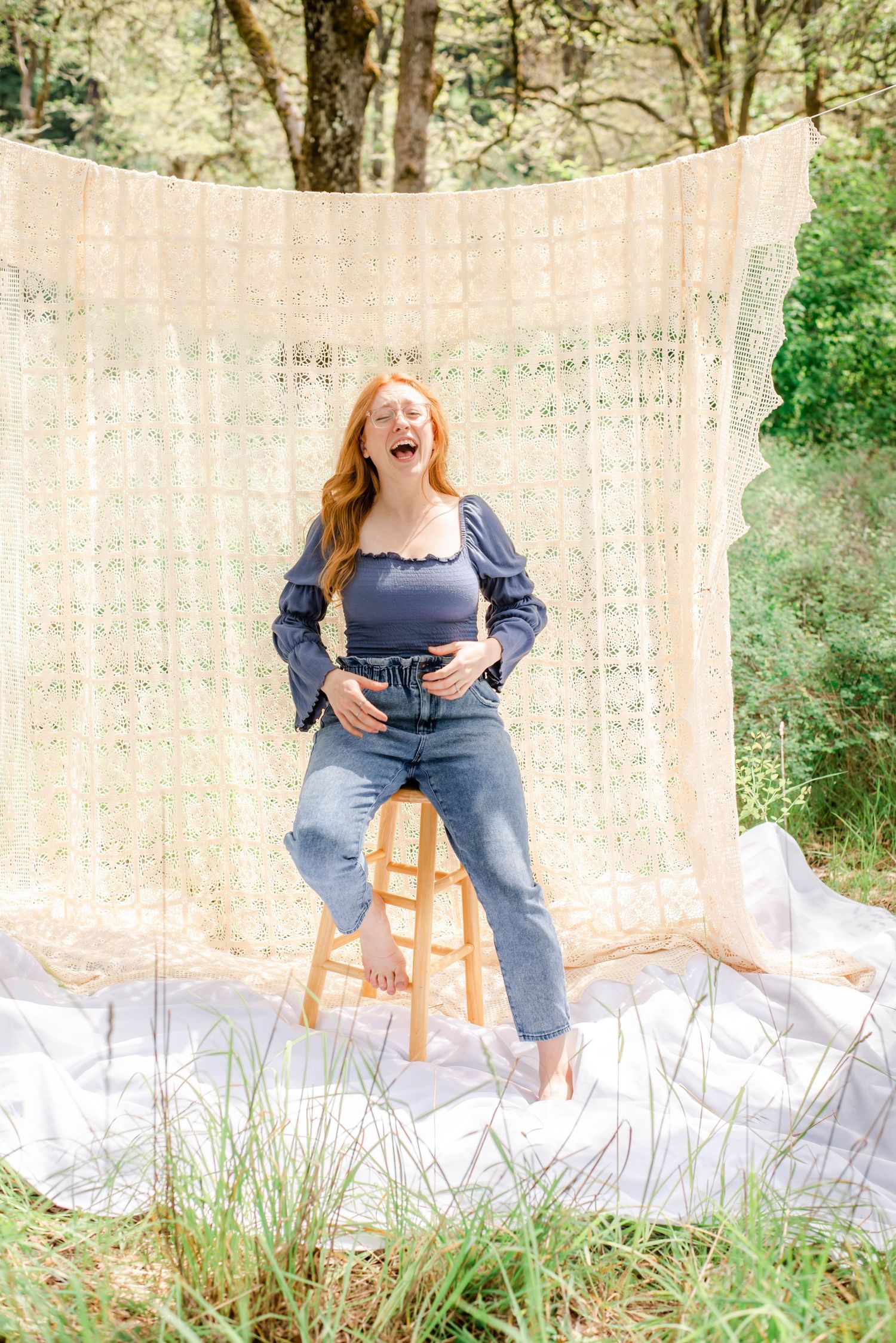 Woman in blue puff sleeve top and jeans poses playfully on wooden stool against lace backdrop.