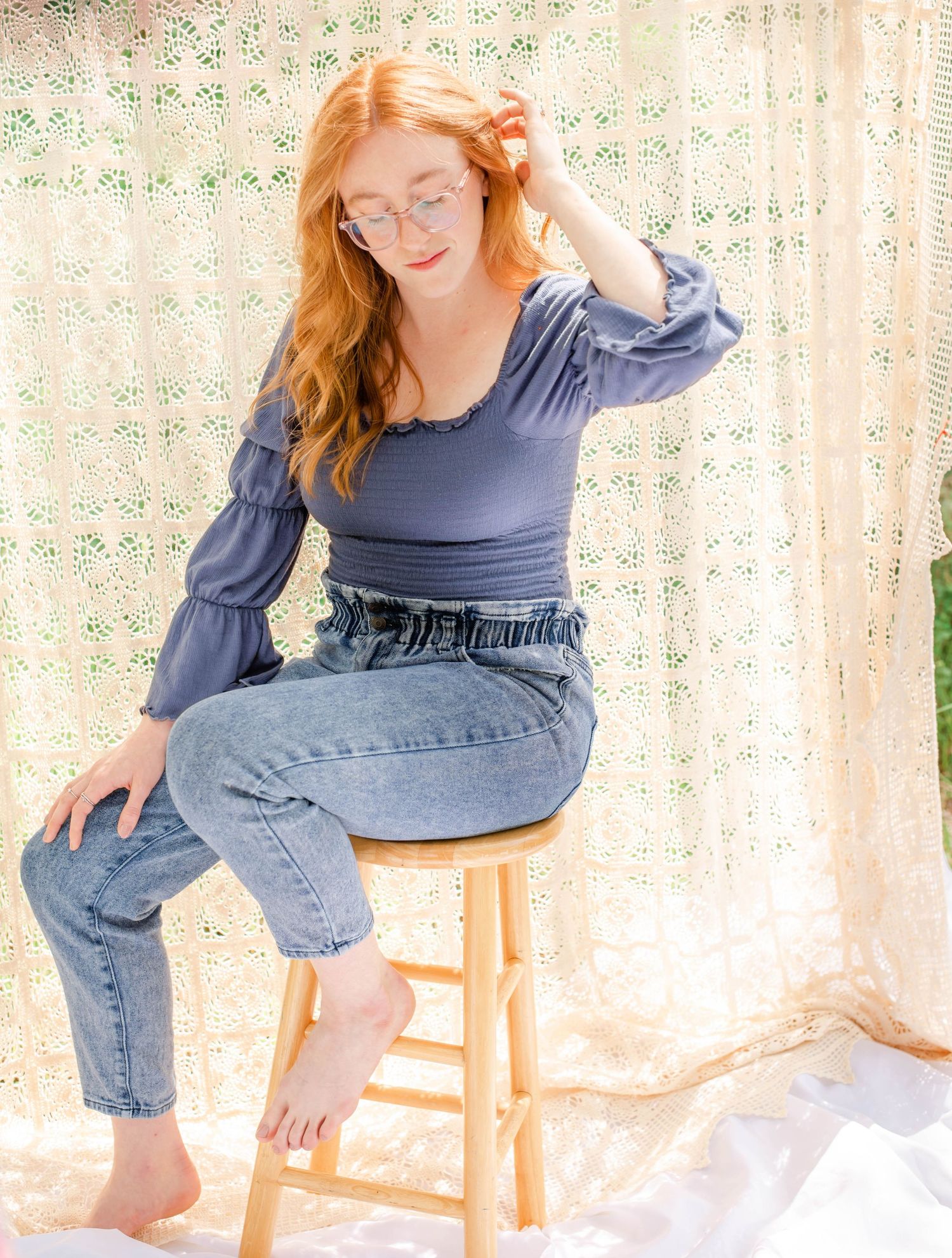 Candid outdoor pose on wooden stool wearing blue top and jeans against delicate lace background.