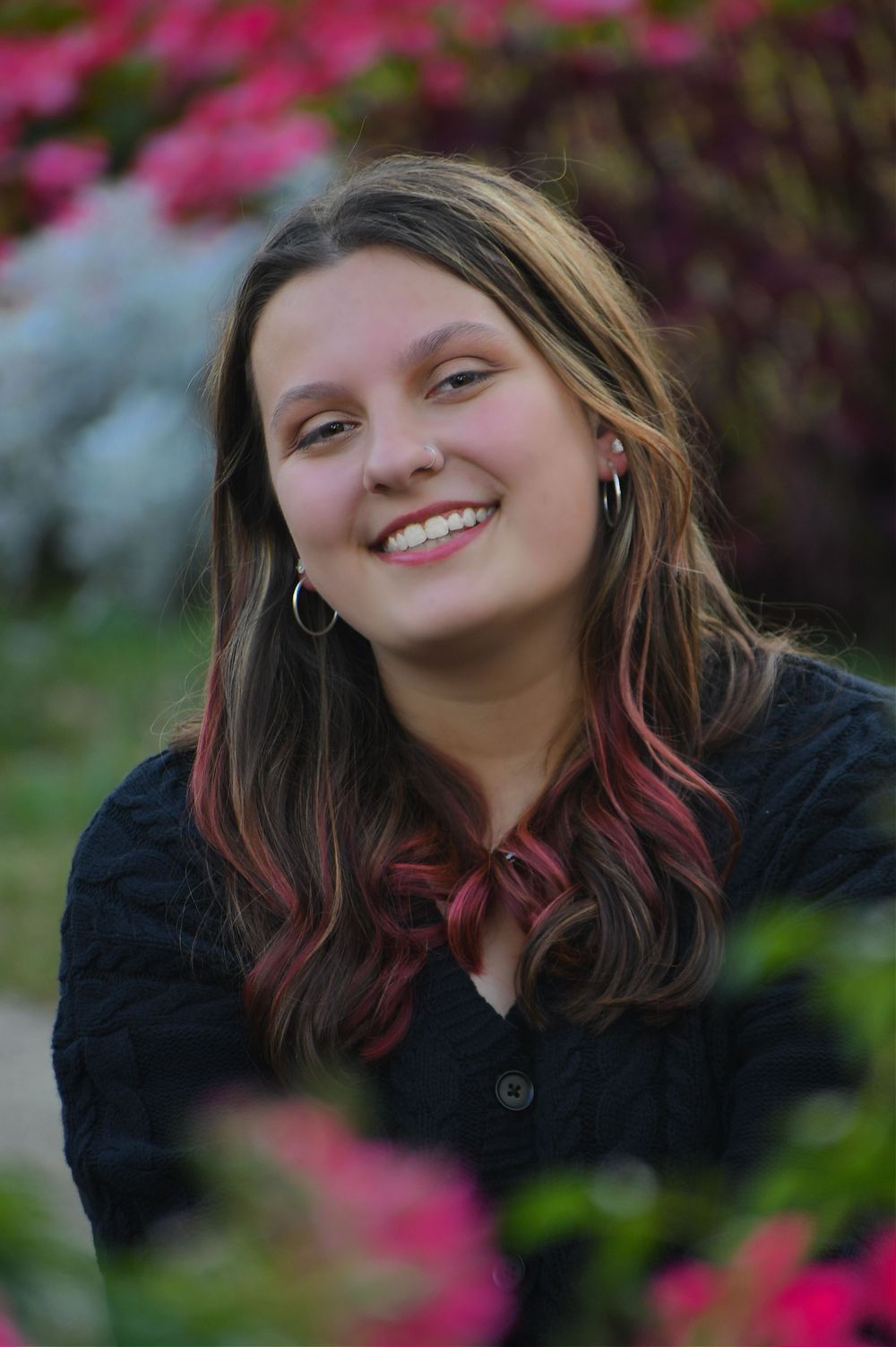 Portrait among pink flowers featuring long hair with colored streaks and black button-up top.