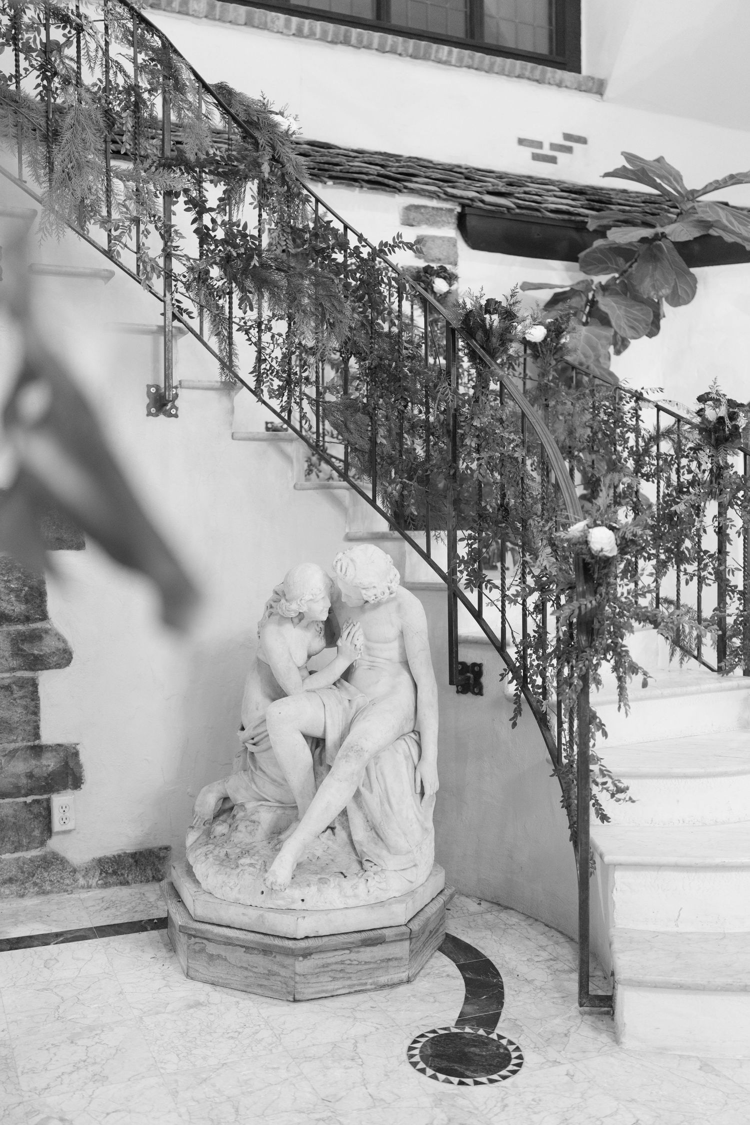 Black and white photo of a classical marble sculpture of two figures beneath an ornate iron staircase adorned with flowers.