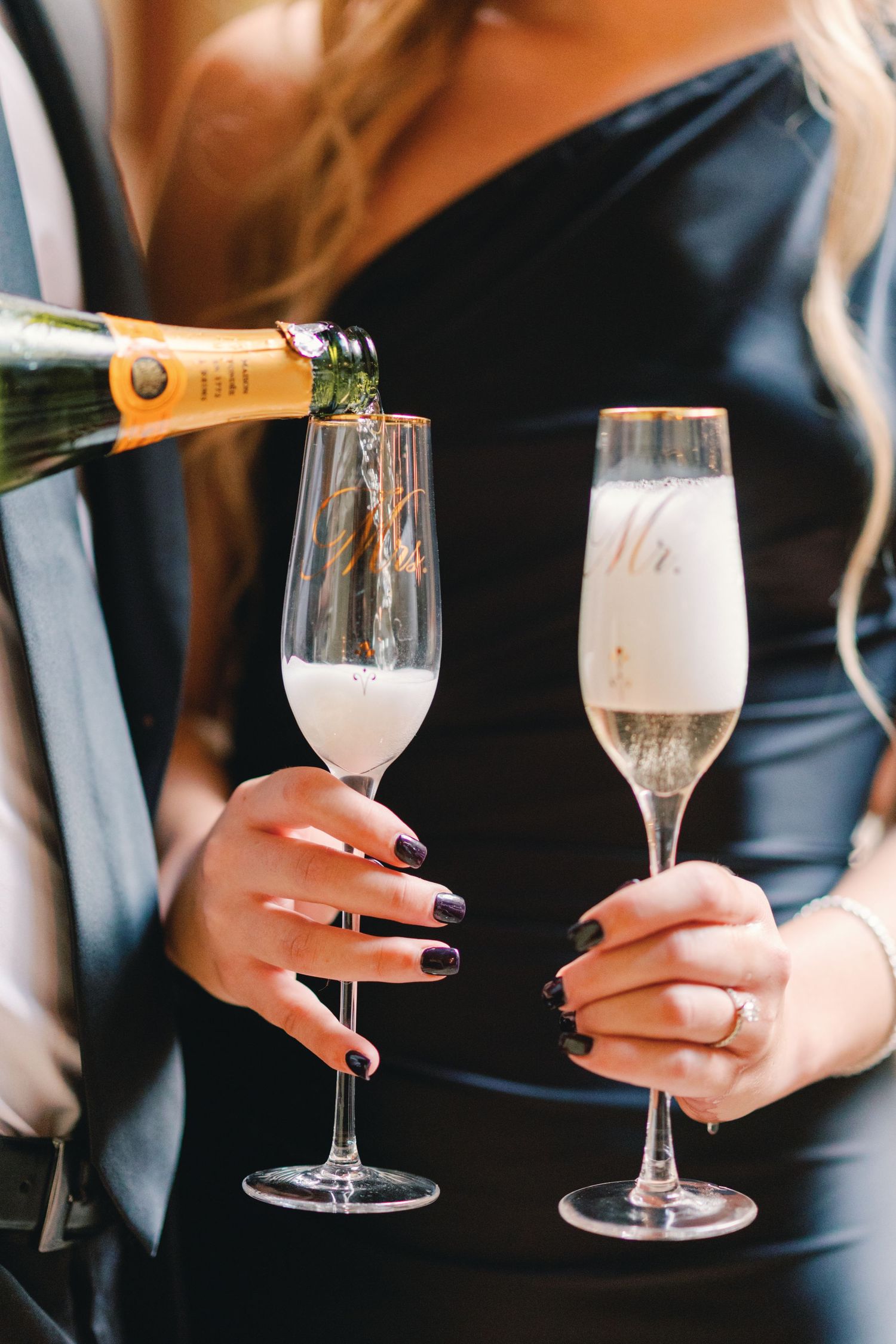 Champagne being poured into Mr. and Mrs. flutes held by bride in black strapless dress.