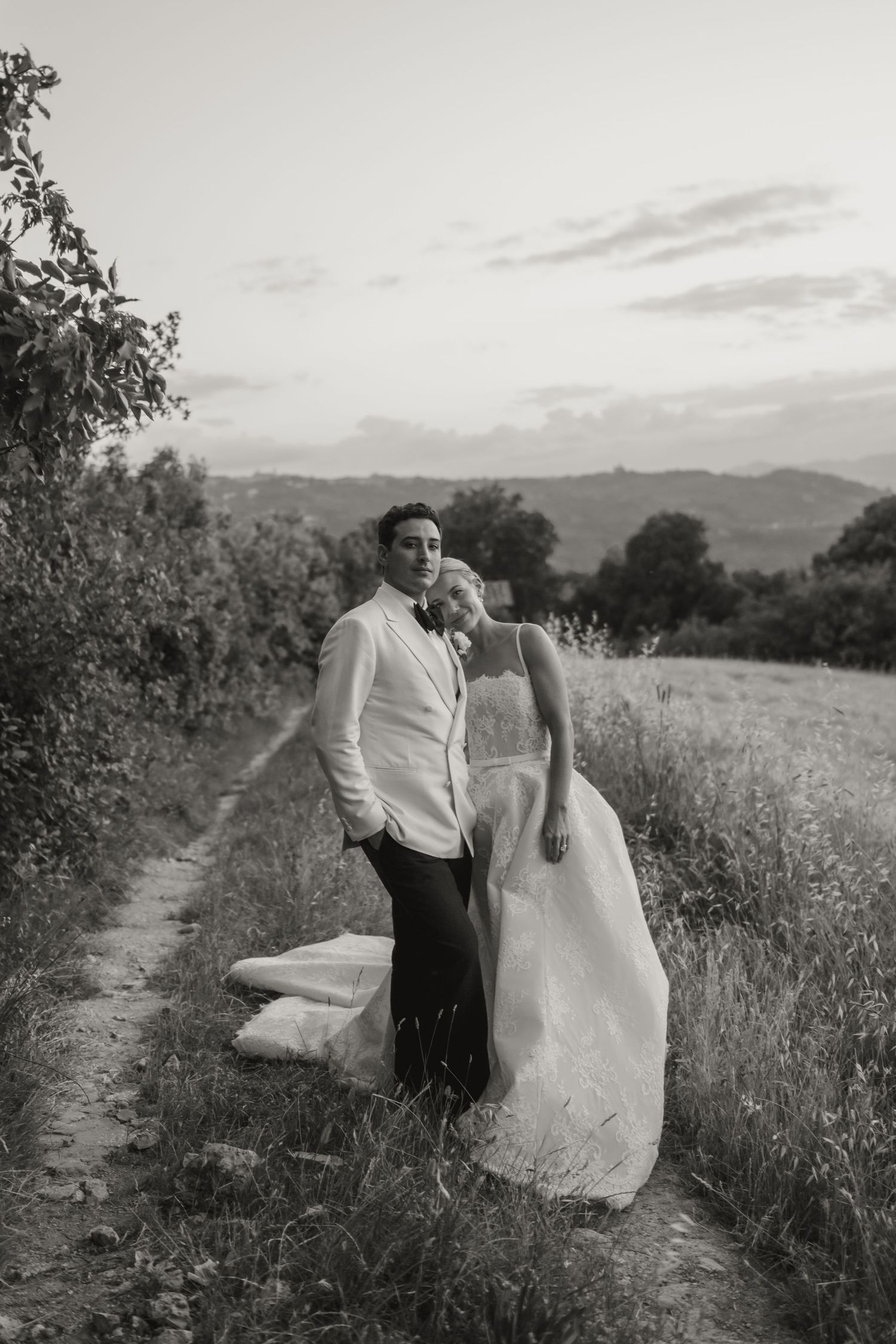 Black and white photo of a bride and groom embracing on a rural path surrounded by lush greenery and rolling hills.