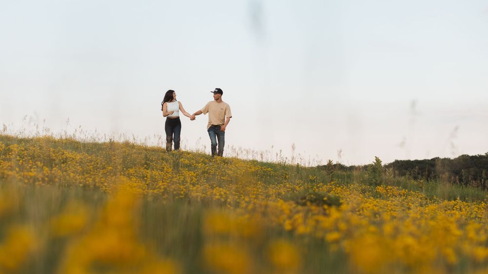 A couple holds hands while walking through a vibrant yellow wildflower field under a bright sky.