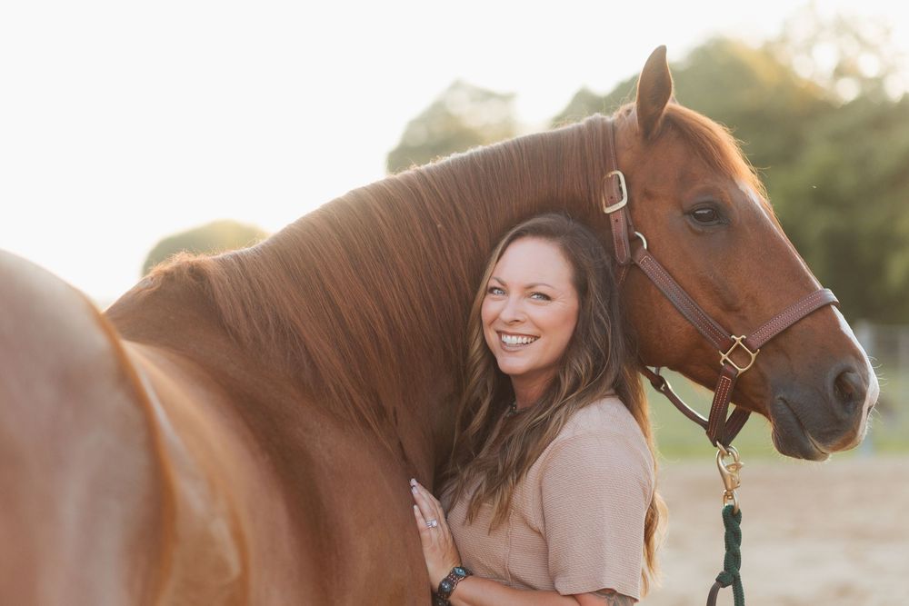 Smiling woman with long brown hair embracing a chestnut horse outdoors in warm golden sunlight.