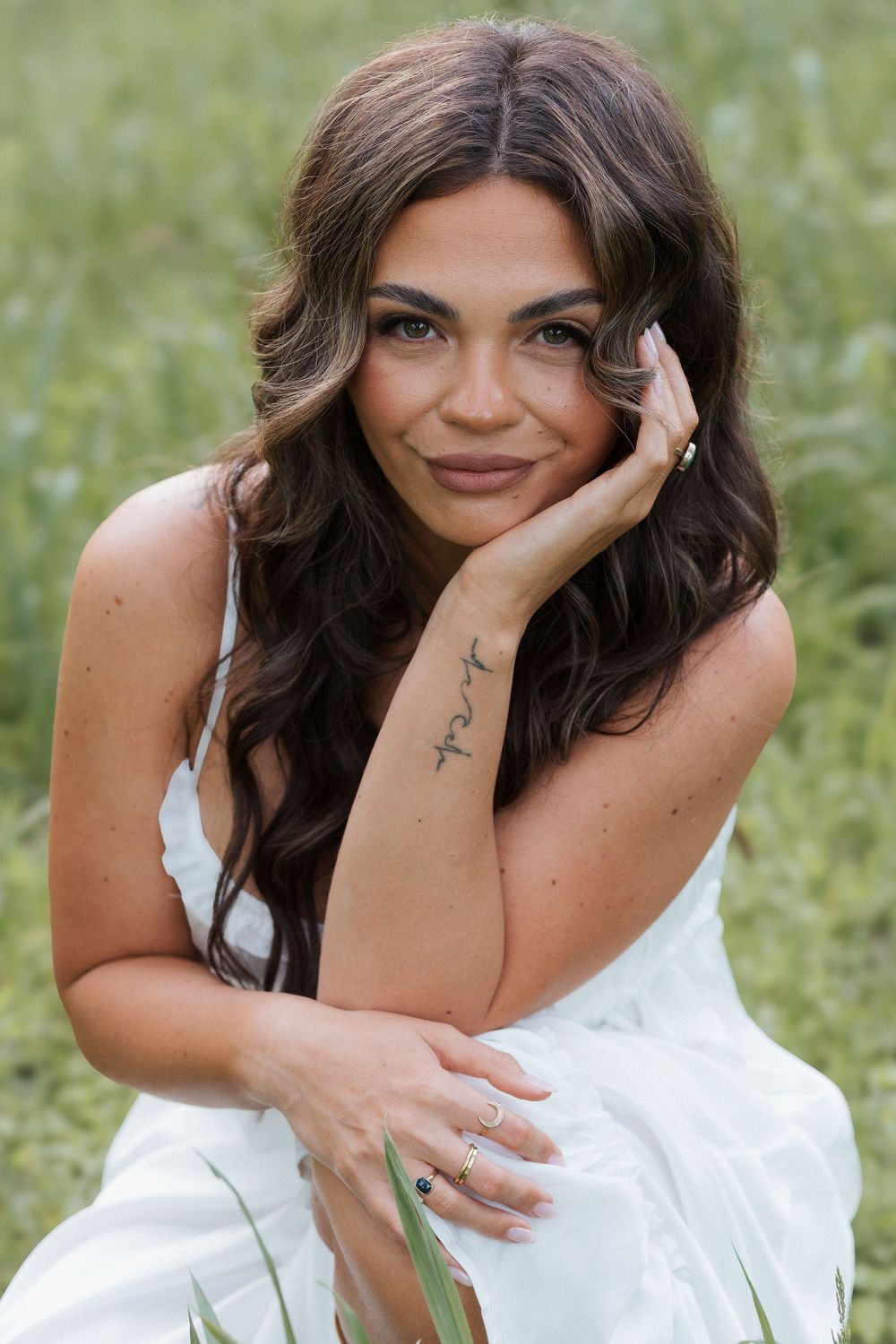 A brunette woman in a white dress sits in a green field, resting her cheek on her hand, showcasing a forearm tattoo.