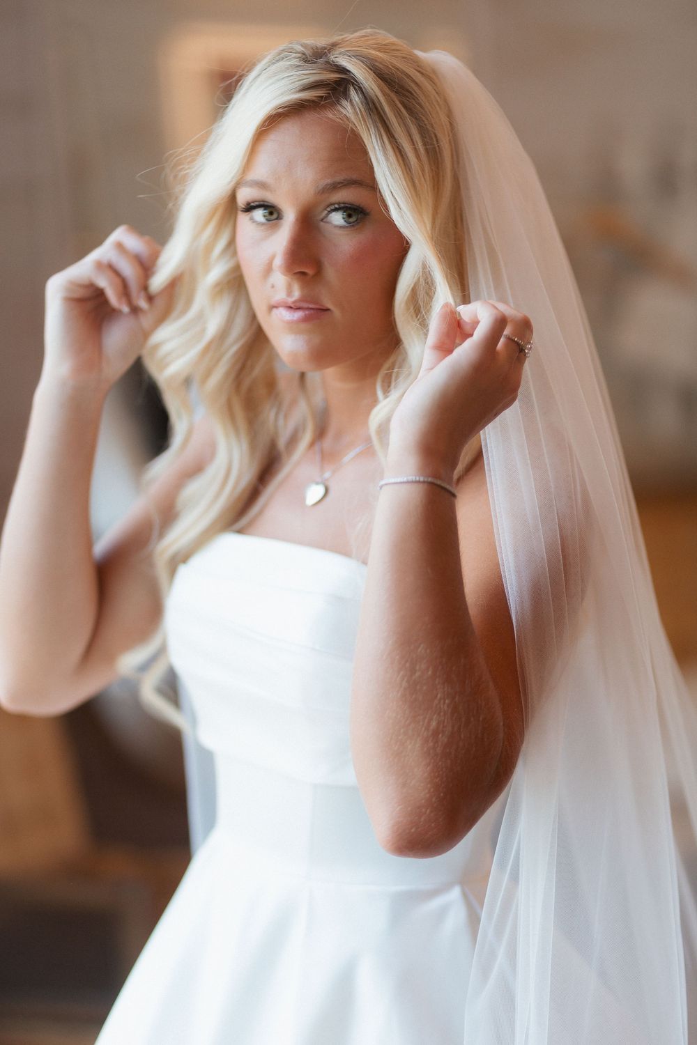 Blonde bride in white strapless gown and veil adjusting her hair, wearing a heart necklace and bracelet.