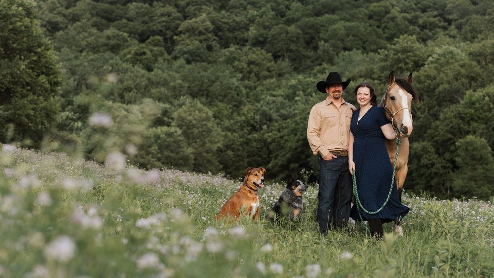 Couple in western wear posing with a horse and two dogs in a lush green meadow with wildflowers.