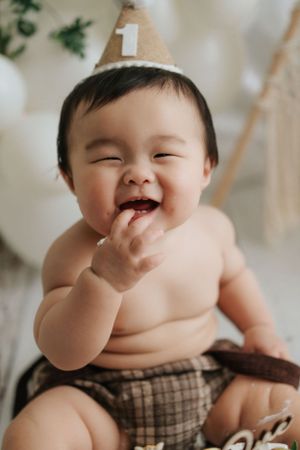 Chubby baby in plaid diaper laughing with birthday hat during milestone photo session.穿著格子尿布、戴著生日帽的胖乎乎的寶寶在里程碑照片拍攝中開懷大笑。