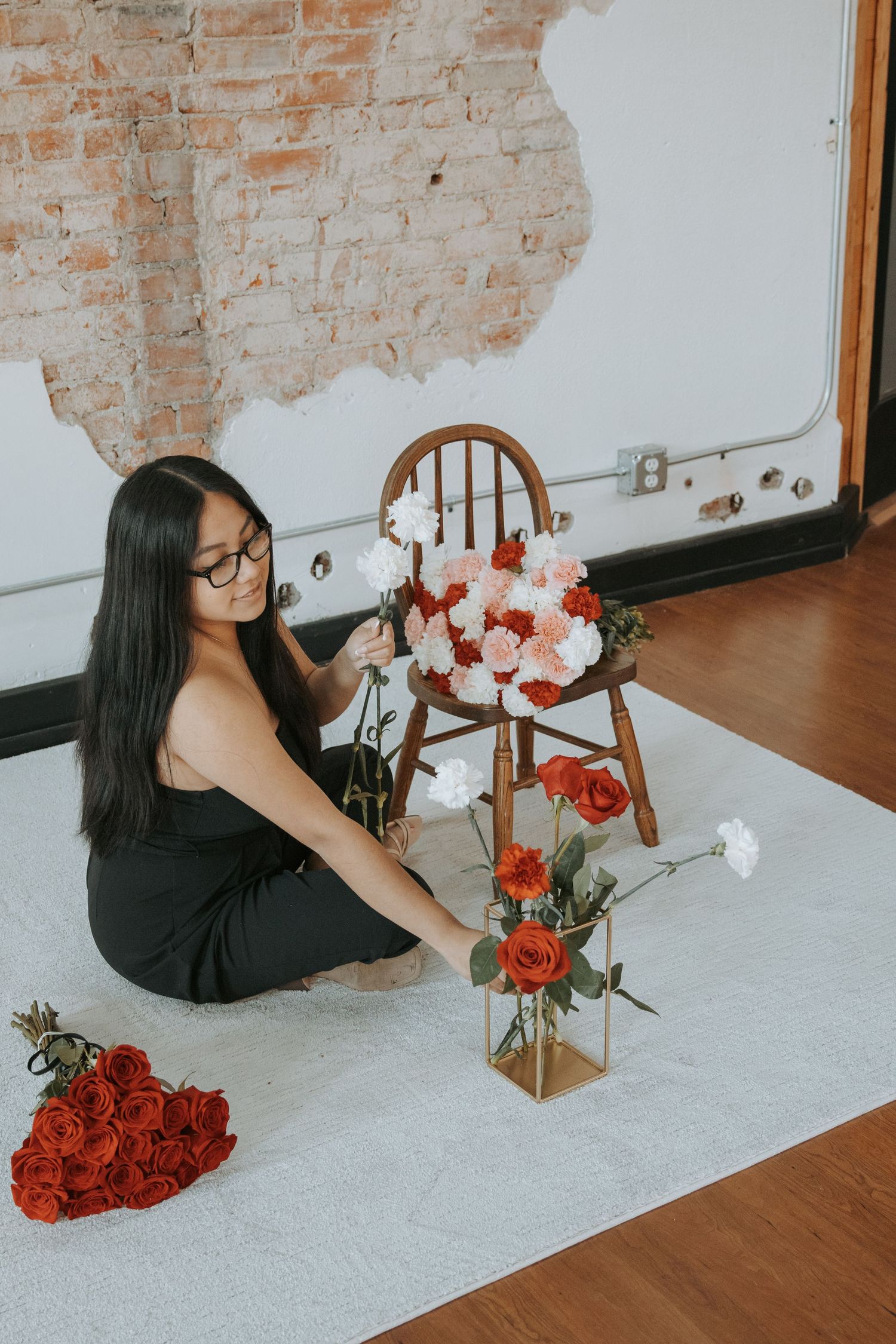 Woman in black dress arranging red and white roses near a wooden chair on a white rug in a studio.