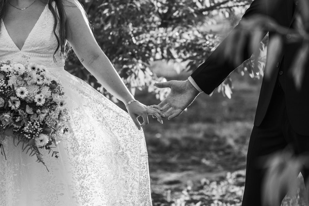 Black and white photo of bride and groom holding hands, bride holding floral bouquet in lace wedding dress.