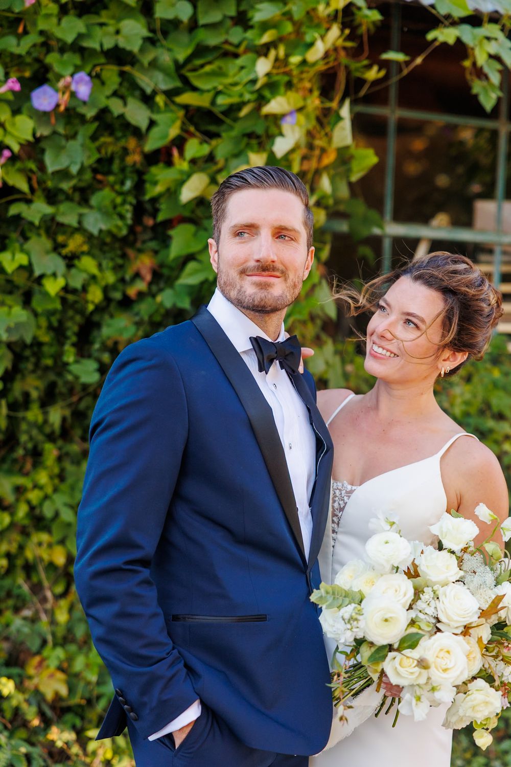 Bride holding white floral bouquet smiles at groom in navy tuxedo standing before lush green ivy wall.
