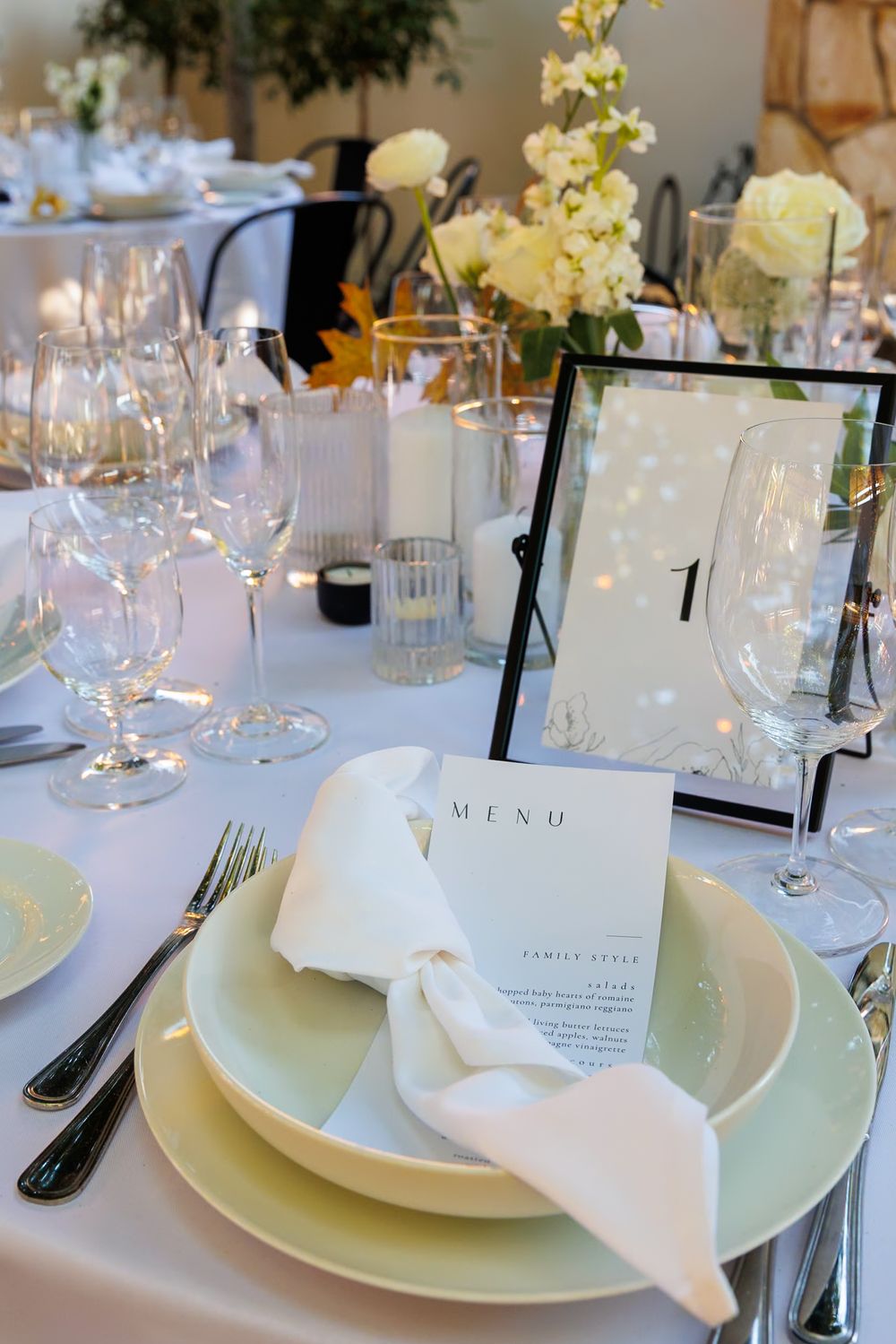 Elegant wedding table setting with white menu card, sage green plates, crystal glasses, and floral centerpiece with table number one.