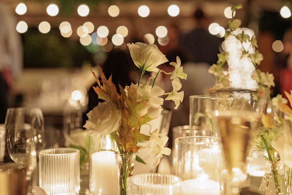 Elegant wedding reception table with white floral centerpiece, candles, and glowing string lights in background.