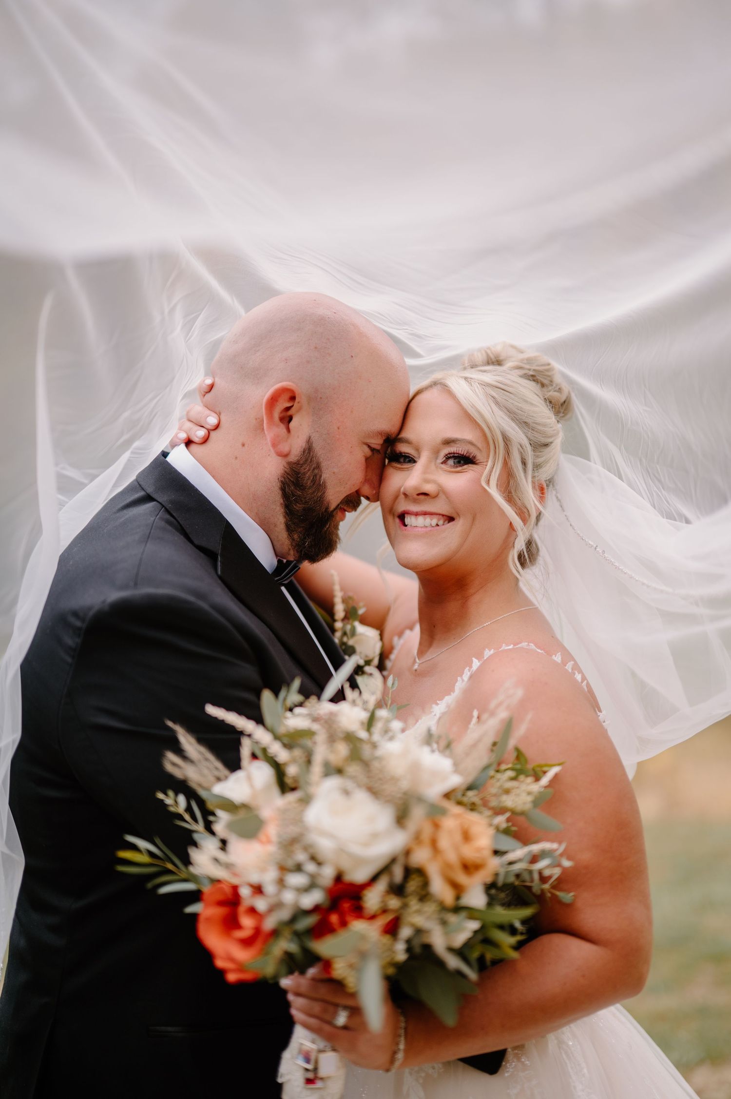 Bride and groom share romantic moment under flowing veil, bride holding colorful bouquet of white, orange, and red flowers.