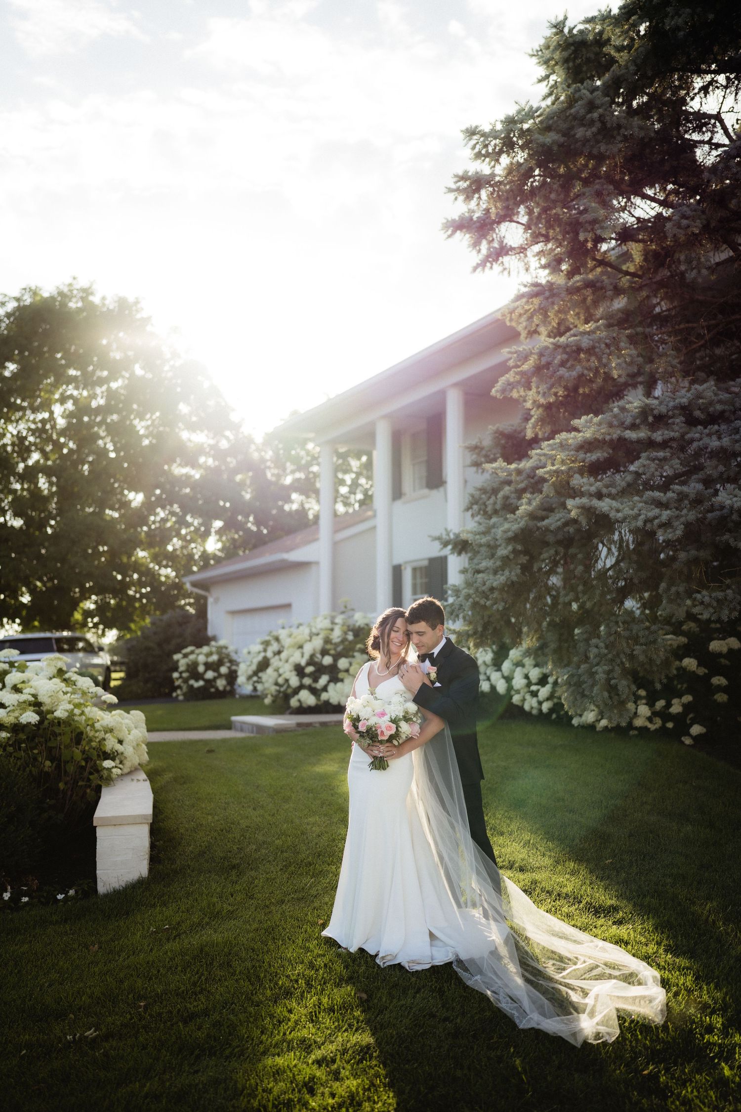 Bride and groom embrace on lush green lawn beside blooming white hydrangeas and elegant white estate at golden hour.