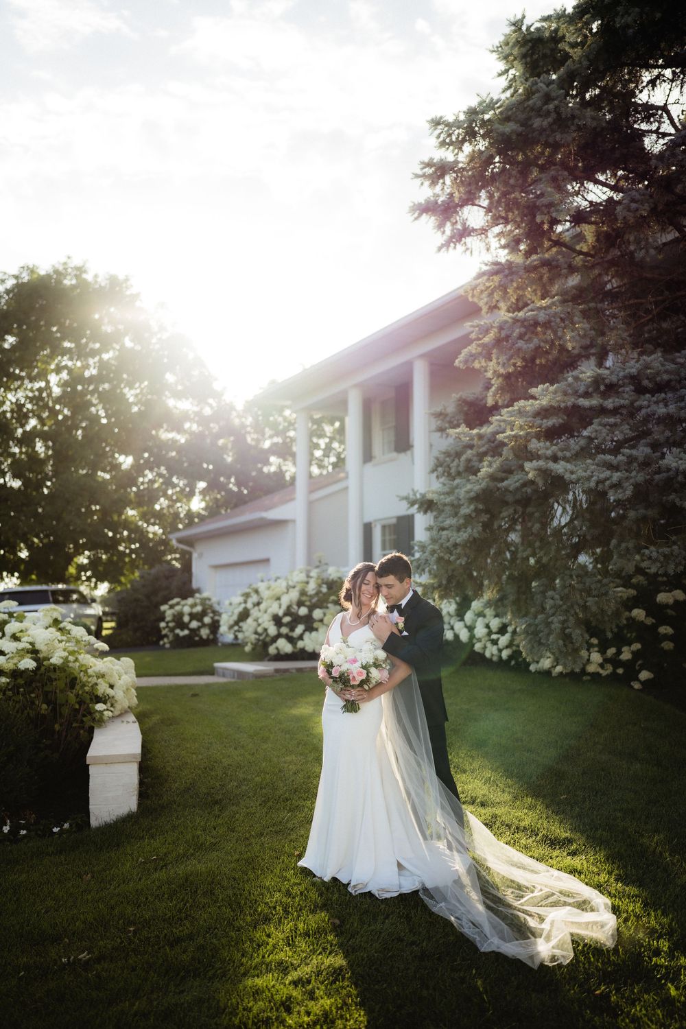 Bride and groom embrace on lush green lawn beside blooming white hydrangeas and elegant white estate at golden hour.