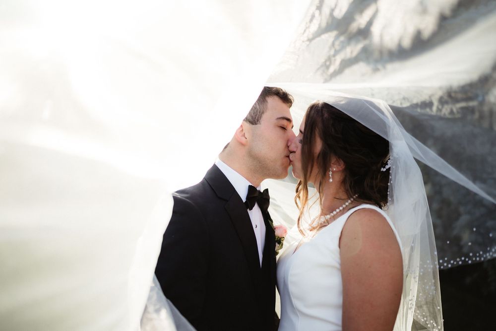 Bride and groom sharing a romantic kiss under a flowing white veil on their wedding day.