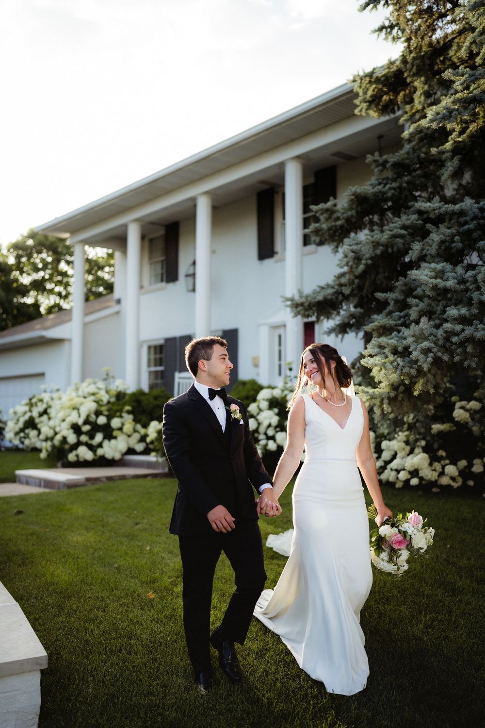 Bride and groom hold hands walking on green lawn before white colonial home with hydrangeas.