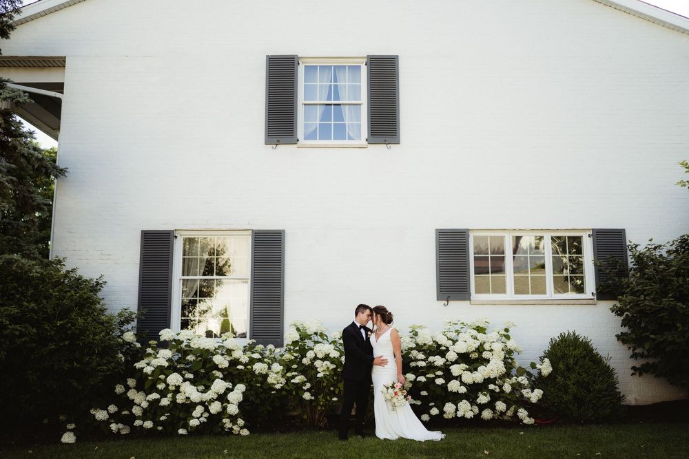Bride and groom kissing in front of white farmhouse with dark shutters and white hydrangeas at outdoor wedding.