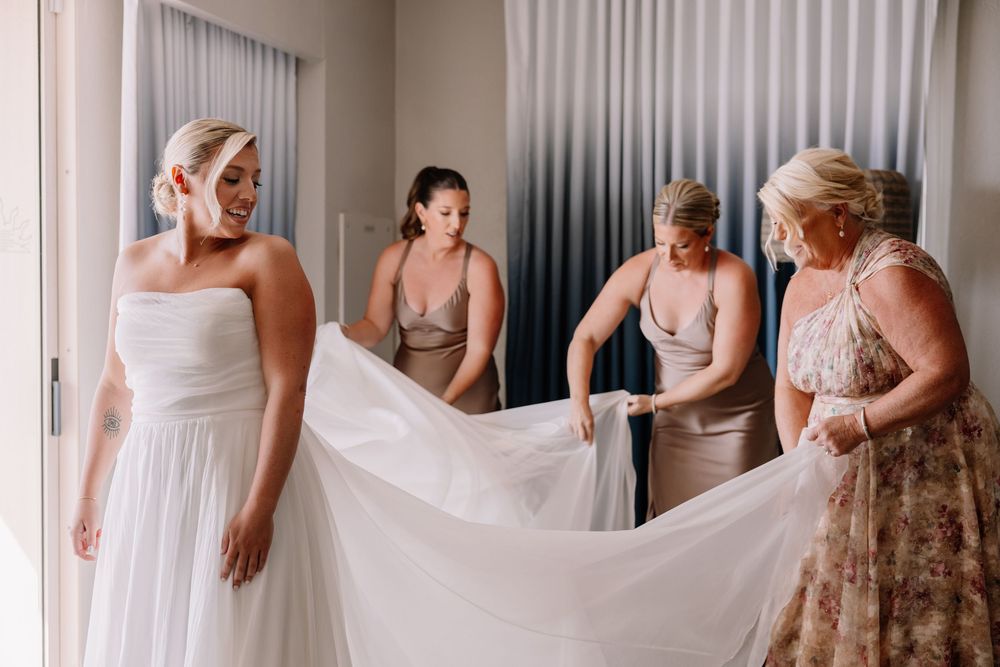 Bridesmaids helping bride with wedding dress train in bright room before ceremony.