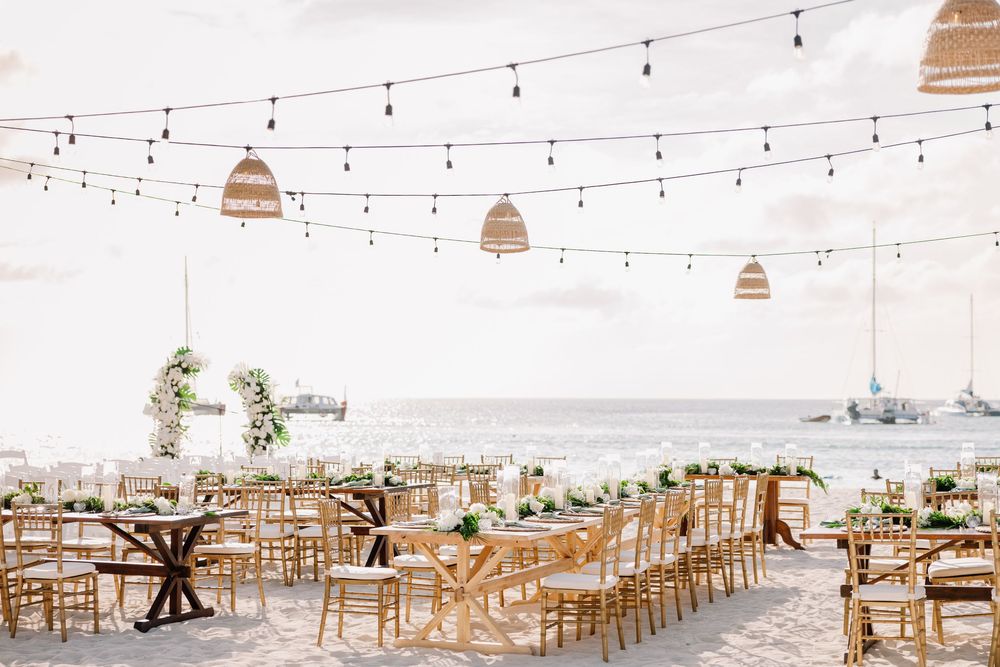 Elegant beachside wedding reception with string lights, wicker pendants, and gold chiavari chairs overlooking the ocean.