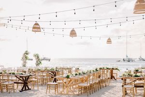 Elegant beachside wedding reception with string lights, wicker pendants, and gold chiavari chairs overlooking the ocean.
