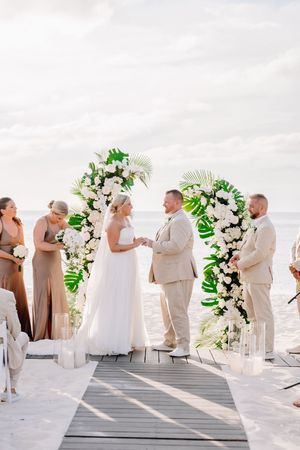 Bride and groom exchange vows at a tropical beach wedding under a floral arch with white blooms and palm leaves.