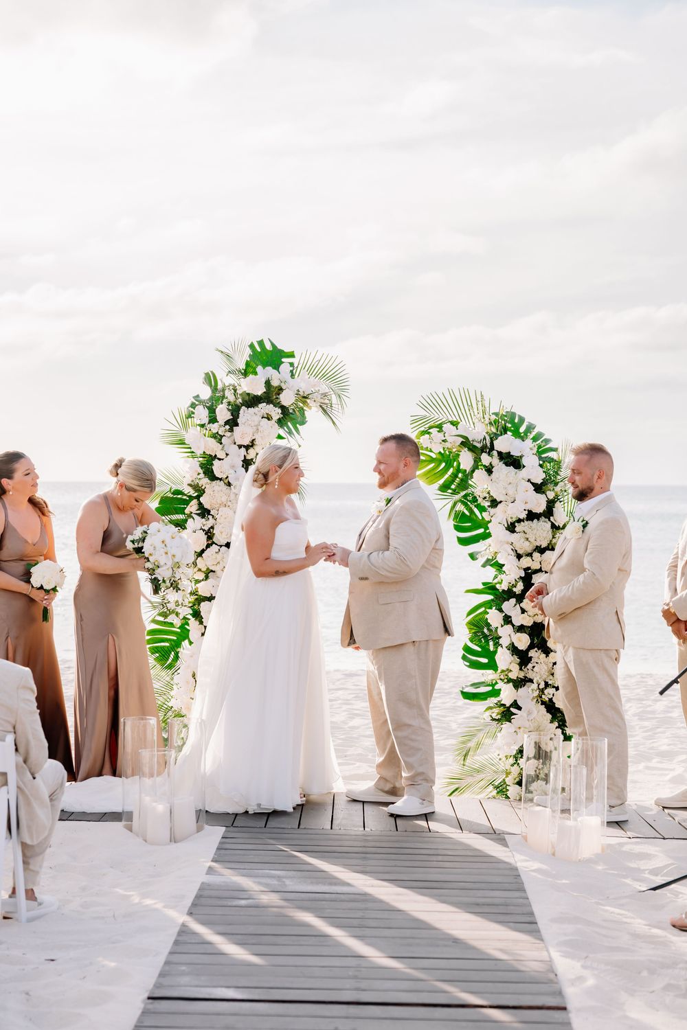 Bride and groom exchange vows at a tropical beach wedding under a floral arch with white blooms and palm leaves.