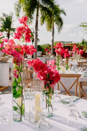 Elegant outdoor wedding reception tables with vibrant pink tropical flowers in glass vases, white candles, and palm trees in background.