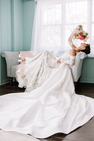 Bride in lace long-sleeve wedding gown with train reclines on sofa, holding floral bouquet aloft near bright window.