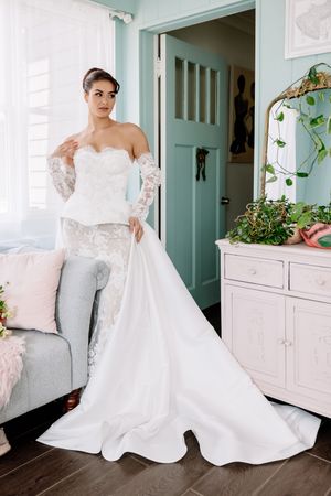 Bride in strapless lace wedding gown with long train and lace gloves posing in elegant pastel room.